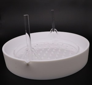 A white circular ceramic container with a spout on the side and a textured base is placed against a dark background. Two tall, clear plastic rods are inserted vertically into the container, creating a symmetrical structure.