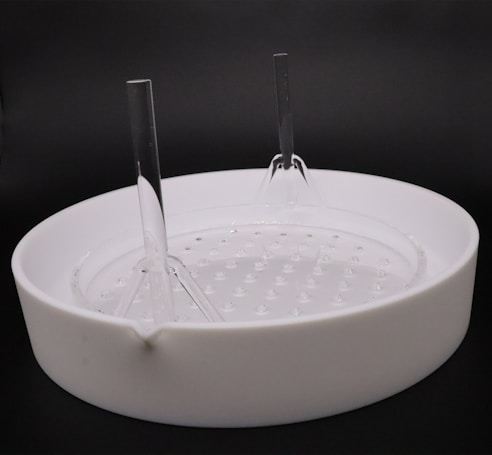A white circular ceramic container with a spout on the side and a textured base is placed against a dark background. Two tall, clear plastic rods are inserted vertically into the container, creating a symmetrical structure.