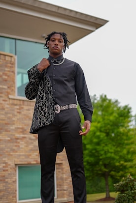 A person is standing confidently in an outdoor setting, wearing a dark, stylish outfit with a patterned jacket draped over one shoulder. The background features a modern building with large windows and a greenery-filled landscape.