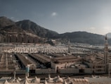 A vast array of white tents is spread across a large open area surrounded by mountainous terrain. Two minarets stand prominently in the foreground. The background consists of rugged hills under a clear sky, and there are several uniform, rectangular buildings at the base of the mountains.