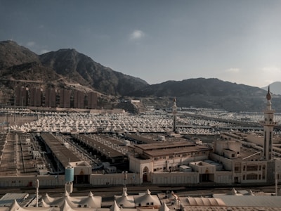 A vast array of white tents is spread across a large open area surrounded by mountainous terrain. Two minarets stand prominently in the foreground. The background consists of rugged hills under a clear sky, and there are several uniform, rectangular buildings at the base of the mountains.