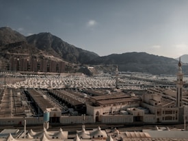 A vast array of white tents is spread across a large open area surrounded by mountainous terrain. Two minarets stand prominently in the foreground. The background consists of rugged hills under a clear sky, and there are several uniform, rectangular buildings at the base of the mountains.
