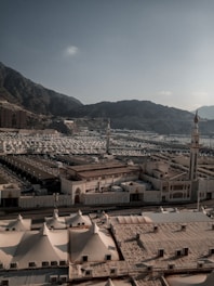 A vast landscape filled with numerous white tents arranged in an orderly manner. The tents are surrounded by a mountainous backdrop under a clear sky. There are several taller structures, possibly minarets, scattered among the tents, contributing to an organized city-like pattern.