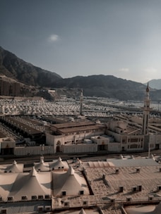 A vast landscape filled with numerous white tents arranged in an orderly manner. The tents are surrounded by a mountainous backdrop under a clear sky. There are several taller structures, possibly minarets, scattered among the tents, contributing to an organized city-like pattern.