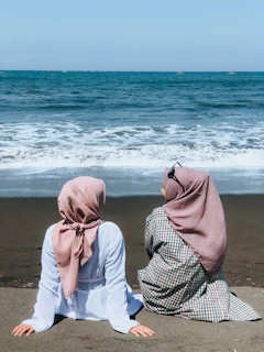 two women sitting on a beach facing the ocean