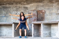 A joyful moment between two people is captured as one sits on the other's back with arms outstretched, giving the appearance of flight. They stand within a concrete structure with visible graffiti and metallic elements, suggesting an urban or industrial setting.