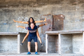 A joyful moment between two people is captured as one sits on the other's back with arms outstretched, giving the appearance of flight. They stand within a concrete structure with visible graffiti and metallic elements, suggesting an urban or industrial setting.