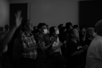 A group of people are standing together in an indoor setting, appearing to be in a moment of attention or prayer. Some individuals are wearing masks and have their eyes closed, with their hands raised or clasped. The image has a grayscale color palette.