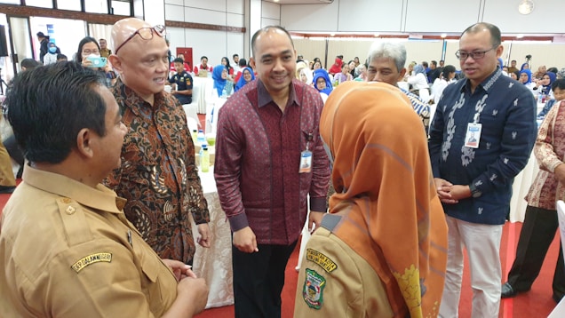 A group of people is gathered in a well-lit conference room, dressed in formal and semi-formal attire. One woman is wearing a headscarf and a uniform with an official insignia. They appear to be engaging in a conversation, surrounded by tables and chairs with other attendees in the background.