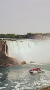 A massive waterfall cascades powerfully over a cliff, surrounded by lush greenery. A tour boat full of passengers wearing red ponchos navigates the misty waters at the base of the falls. In the background, a large building is visible near the wooded shoreline.