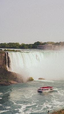 A massive waterfall cascades powerfully over a cliff, surrounded by lush greenery. A tour boat full of passengers wearing red ponchos navigates the misty waters at the base of the falls. In the background, a large building is visible near the wooded shoreline.