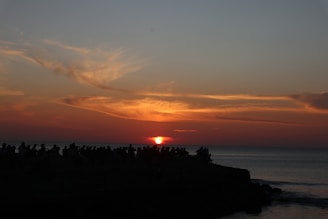 The image captures a breathtaking sunset over the ocean, with vibrant orange and red hues blending into the darkening sky. Silhouettes of people are visible along the shoreline, gathered to witness the sun setting on the horizon.