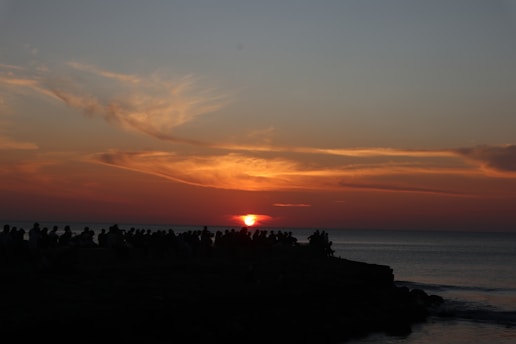 The image captures a breathtaking sunset over the ocean, with vibrant orange and red hues blending into the darkening sky. Silhouettes of people are visible along the shoreline, gathered to witness the sun setting on the horizon.