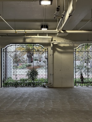 An industrial-style interior space with decorative metal grilles in front of large windows. Exposed pipes on the ceiling and walls add to the industrial aesthetic. The floor appears to be concrete, and outside the windows, vegetation is visible.