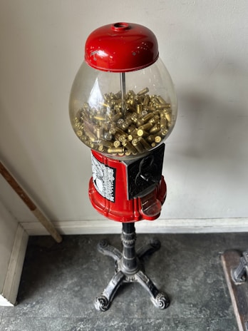 A vintage gumball machine with a red top and black metal stand is filled with brass bullet casings instead of gumballs. It is placed against a plain wall on a dark-colored floor.