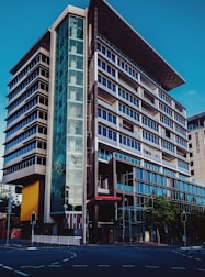 A modern multi-story office building with a sleek and architectural design featuring prominent glass facades and a unique angular rooftop. The structure has a combination of dark and light hues, with a significant glass entrance on the corner. Traffic lights and a city street are visible in the foreground.