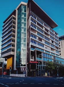 A modern multi-story office building with a sleek and architectural design featuring prominent glass facades and a unique angular rooftop. The structure has a combination of dark and light hues, with a significant glass entrance on the corner. Traffic lights and a city street are visible in the foreground.