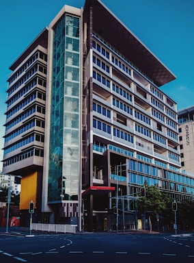 A modern multi-story office building with a sleek and architectural design featuring prominent glass facades and a unique angular rooftop. The structure has a combination of dark and light hues, with a significant glass entrance on the corner. Traffic lights and a city street are visible in the foreground.