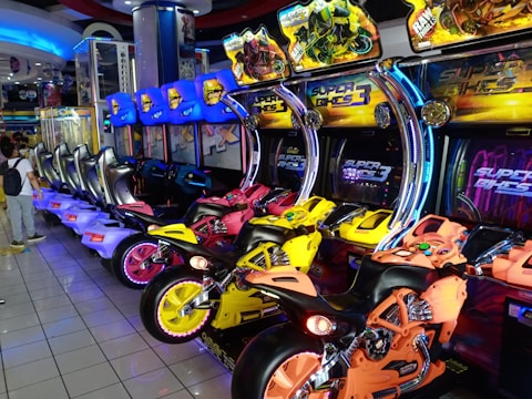 An arcade setting featuring a row of colorful motorcycle racing arcade games, each equipped with bright neon lights and vibrant designs. The motorcycles are in various colors including yellow, orange, and pink. Monitors display the game title 'Super Bikes 3' above each motorcycle. The room is well-lit, with reflective tiled flooring and a person standing nearby, adding to the lively atmosphere.