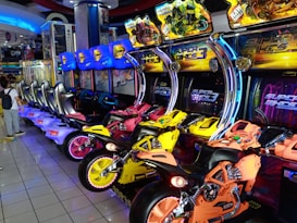 An arcade setting featuring a row of colorful motorcycle racing arcade games, each equipped with bright neon lights and vibrant designs. The motorcycles are in various colors including yellow, orange, and pink. Monitors display the game title 'Super Bikes 3' above each motorcycle. The room is well-lit, with reflective tiled flooring and a person standing nearby, adding to the lively atmosphere.
