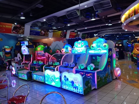 A brightly lit arcade filled with colorful gaming machines sits under ceiling lights. Neon lights illuminate various game stations, creating a playful and vibrant environment. A few red chairs are scattered around the reflective, tiled floor, and a person is observing one of the machines. The atmosphere is lively and cheerful, featuring games with cartoonish designs and engaging themes.