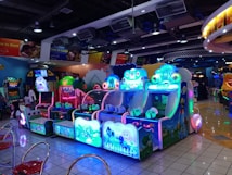 A brightly lit arcade filled with colorful gaming machines sits under ceiling lights. Neon lights illuminate various game stations, creating a playful and vibrant environment. A few red chairs are scattered around the reflective, tiled floor, and a person is observing one of the machines. The atmosphere is lively and cheerful, featuring games with cartoonish designs and engaging themes.