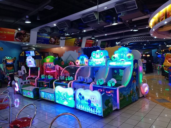 A brightly lit arcade filled with colorful gaming machines sits under ceiling lights. Neon lights illuminate various game stations, creating a playful and vibrant environment. A few red chairs are scattered around the reflective, tiled floor, and a person is observing one of the machines. The atmosphere is lively and cheerful, featuring games with cartoonish designs and engaging themes.