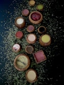 A collection of wooden bowls and containers filled with various powders and substances is arranged on a dark surface scattered with dried herbs. The bowls contain colorful powders including red, white, brown, yellow, and green hues.