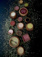 A collection of wooden bowls and containers filled with various powders and substances is arranged on a dark surface scattered with dried herbs. The bowls contain colorful powders including red, white, brown, yellow, and green hues.