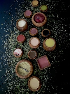 A collection of wooden bowls and containers filled with various powders and substances is arranged on a dark surface scattered with dried herbs. The bowls contain colorful powders including red, white, brown, yellow, and green hues.