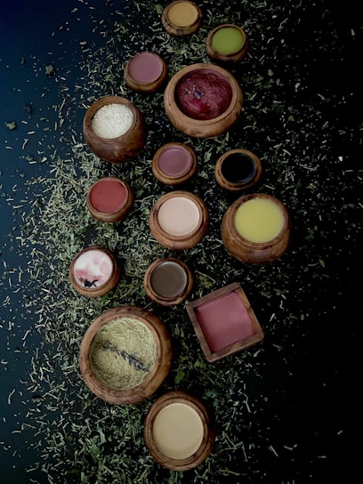 A collection of wooden bowls and containers filled with various powders and substances is arranged on a dark surface scattered with dried herbs. The bowls contain colorful powders including red, white, brown, yellow, and green hues.