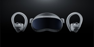 A futuristic-looking virtual reality headset is positioned at the center against a dark background, flanked by two matching controllers on either side. The headset has a sleek, curved visor with a glossy front and appears to have adjustable straps. The controllers feature ergonomic grips and circular designs with buttons and joysticks.