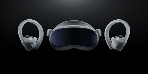 A futuristic-looking virtual reality headset is positioned at the center against a dark background, flanked by two matching controllers on either side. The headset has a sleek, curved visor with a glossy front and appears to have adjustable straps. The controllers feature ergonomic grips and circular designs with buttons and joysticks.