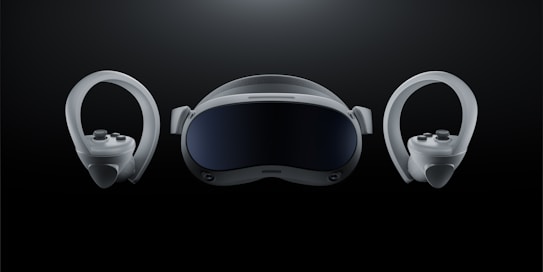 A futuristic-looking virtual reality headset is positioned at the center against a dark background, flanked by two matching controllers on either side. The headset has a sleek, curved visor with a glossy front and appears to have adjustable straps. The controllers feature ergonomic grips and circular designs with buttons and joysticks.