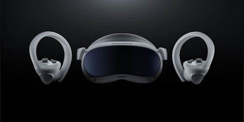 A futuristic-looking virtual reality headset is positioned at the center against a dark background, flanked by two matching controllers on either side. The headset has a sleek, curved visor with a glossy front and appears to have adjustable straps. The controllers feature ergonomic grips and circular designs with buttons and joysticks.
