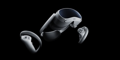 A set of virtual reality equipment, including a headset and two handheld controllers, displayed against a black background. The headset has a sleek, modern design with a glossy surface, while the controllers feature several buttons and joysticks.