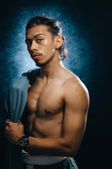 A muscular man with long, dark hair partially tied back, is standing shirtless against a dark, textured background. He holds a blue garment over his shoulder and wears a black wristwatch. His expression is confident and intense.