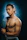 A muscular man with long, dark hair partially tied back, is standing shirtless against a dark, textured background. He holds a blue garment over his shoulder and wears a black wristwatch. His expression is confident and intense.
