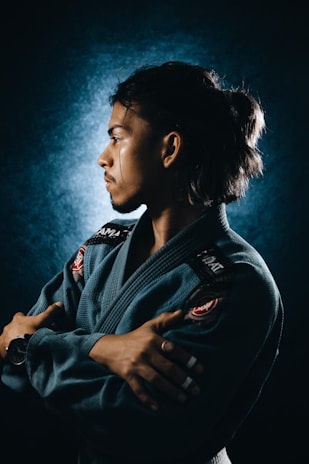 A person wearing a blue martial arts gi, looking to the left with arms crossed. The background is dark, and there is a spotlight effect creating a halo around their head. The person has taped fingers and a watch on the left wrist.