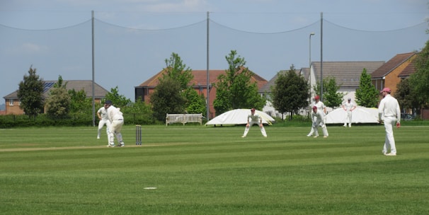 Several people dressed in white uniforms are playing a game of cricket on a well-maintained green field. In the background, there are residential houses with manicured gardens and some trees. The weather appears to be clear with a blue sky.