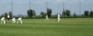 Several players in white uniforms are participating in a cricket match on a grass field. One player is batting while others are fielding. In the background, there are trees, a fence, and the sky is partly cloudy.