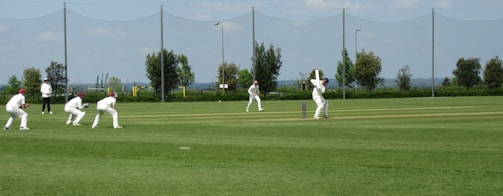 Several players in white uniforms are participating in a cricket match on a grass field. One player is batting while others are fielding. In the background, there are trees, a fence, and the sky is partly cloudy.
