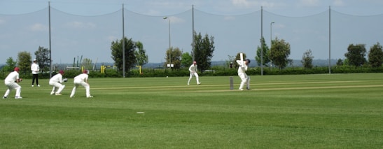 Several players in white uniforms are participating in a cricket match on a grass field. One player is batting while others are fielding. In the background, there are trees, a fence, and the sky is partly cloudy.