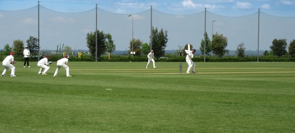A group of people dressed in white cricket uniforms are playing cricket on a well-manicured green field. One player is batting while others, including an umpire, are positioned around him. The background shows a net fence, some trees, and a clear blue sky.