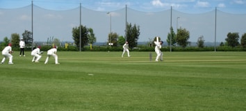 A group of people dressed in white cricket uniforms are playing cricket on a well-manicured green field. One player is batting while others, including an umpire, are positioned around him. The background shows a net fence, some trees, and a clear blue sky.