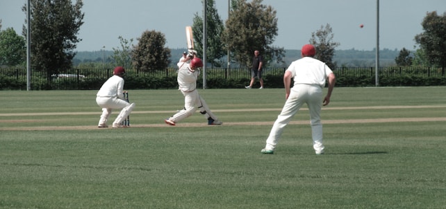 A cricket match scene with players in white uniforms and red caps on a grassy field. One player is mid-swing with a bat, while others are positioned around him, including a wicketkeeper and a bowler in the background. The action captures the moment of a cricket ball in flight.