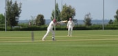 Two people are playing cricket on a well-maintained grassy field. One player is batting while the other is fielding, both wearing traditional white cricket attire and maroon caps. The scene is set outdoors with trees and a distant view of the landscape under a clear sky.