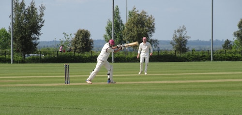 Two people are playing cricket on a well-maintained grassy field. One player is batting while the other is fielding, both wearing traditional white cricket attire and maroon caps. The scene is set outdoors with trees and a distant view of the landscape under a clear sky.