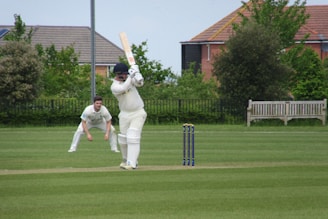 A cricket match is taking place on a grassy field. A batsman in white uniform and helmet is poised to strike the ball with a wooden bat. Behind the batsman, a wicket keeper in a similar white outfit is ready to catch the ball. The scene is set against a backdrop of green bushes and residential houses with visible rooftops. The sky is partly cloudy, adding to the sunny ambiance.