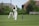 A cricket match is taking place on a grassy field. A batsman in white uniform and helmet is poised to strike the ball with a wooden bat. Behind the batsman, a wicket keeper in a similar white outfit is ready to catch the ball. The scene is set against a backdrop of green bushes and residential houses with visible rooftops. The sky is partly cloudy, adding to the sunny ambiance.